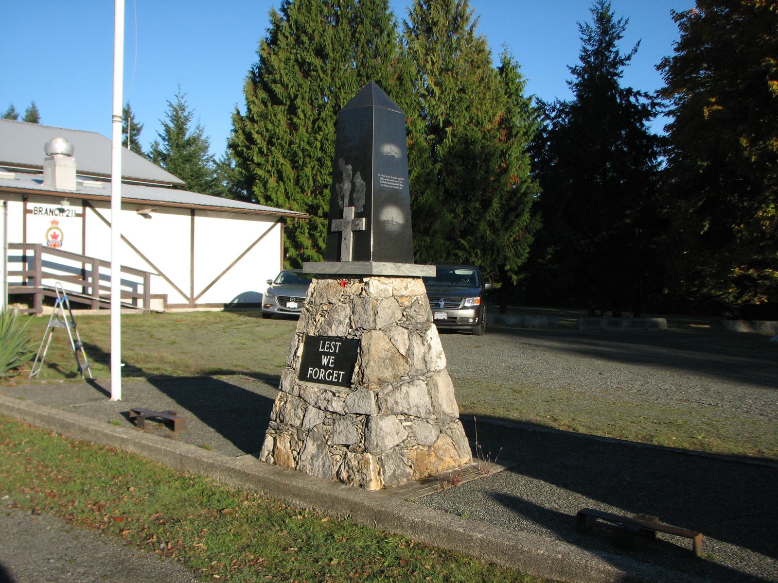 The branch cenotaph: a dark stone obelisk on a fieldstone base with a plaque reading Lest We Forget, beside the flagpole in front of the hall.