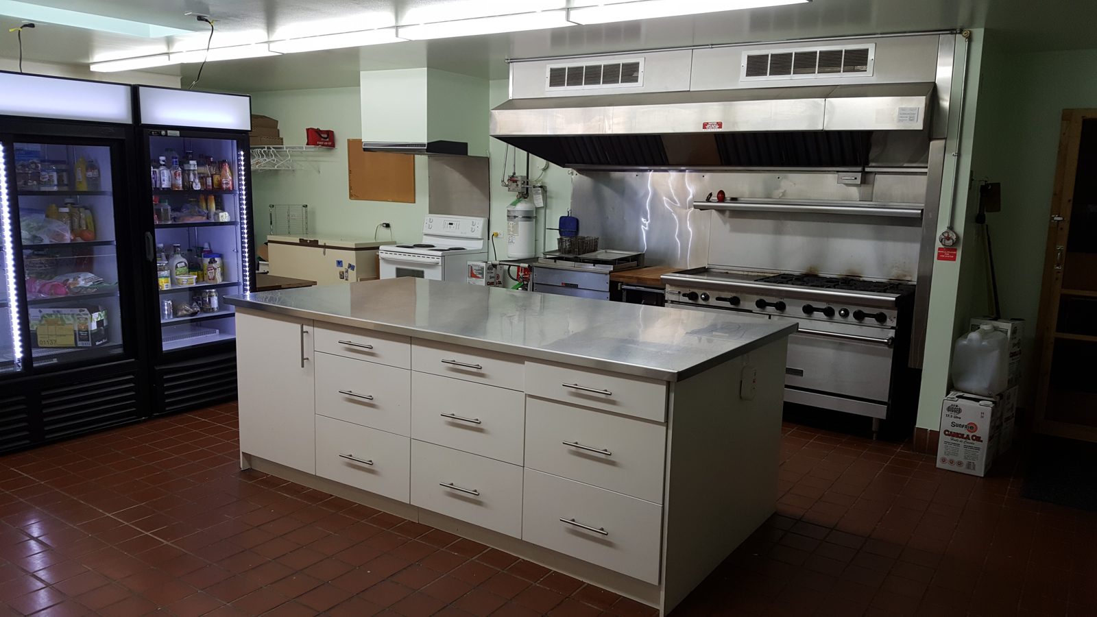 The branch kitchen: a long stainless-steel island, a commercial range under a large hood, and two glass-door fridges.